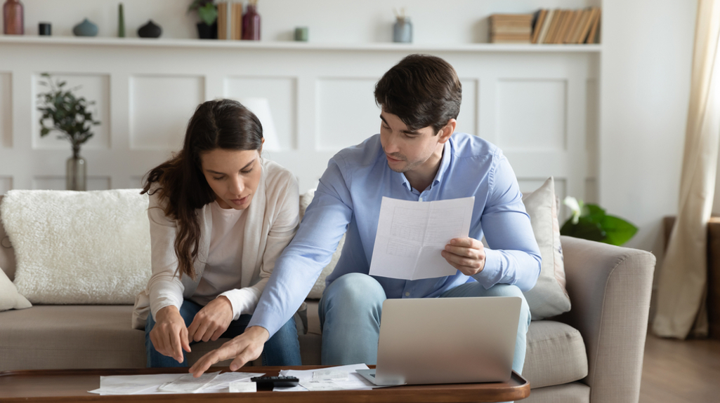 Photo of a couple sat on a sofa looking at papers together on the coffee table infront of them