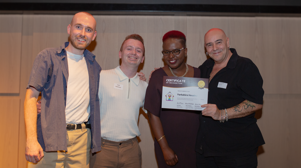 Four people standing together smiling at an event, one of them holding a Certificate of Recognition awarded to Yorkshire Housing.