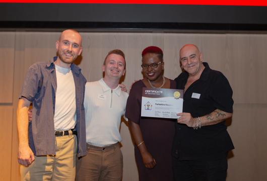 Four people standing together smiling at an event, one of them holding a Certificate of Recognition awarded to Yorkshire Housing.