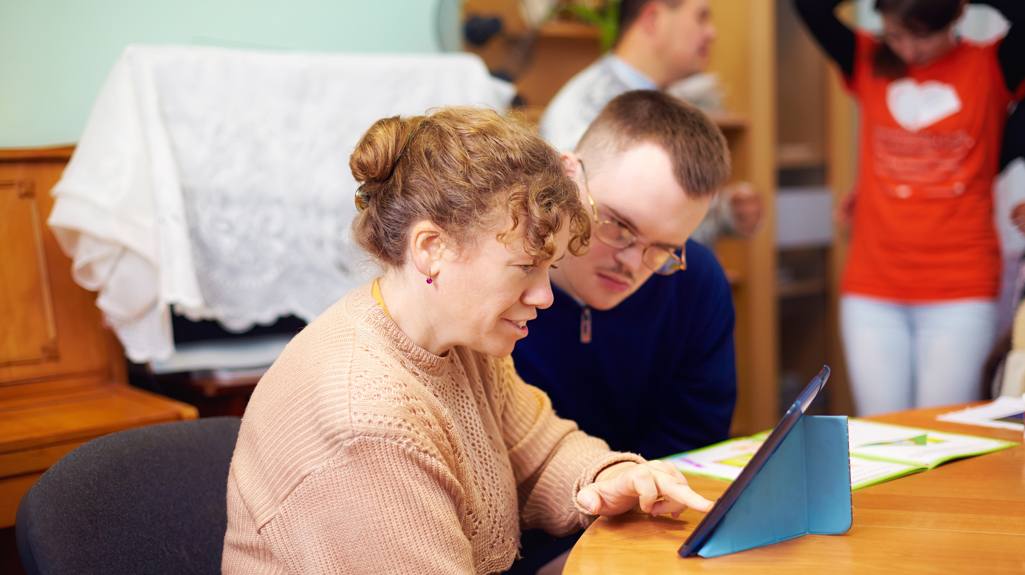 Man and woman sat at a table using an iPad, with one teaching the other how to use it.