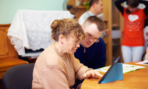 Man and woman sat at a table using an iPad, with one teaching the other how to use it.