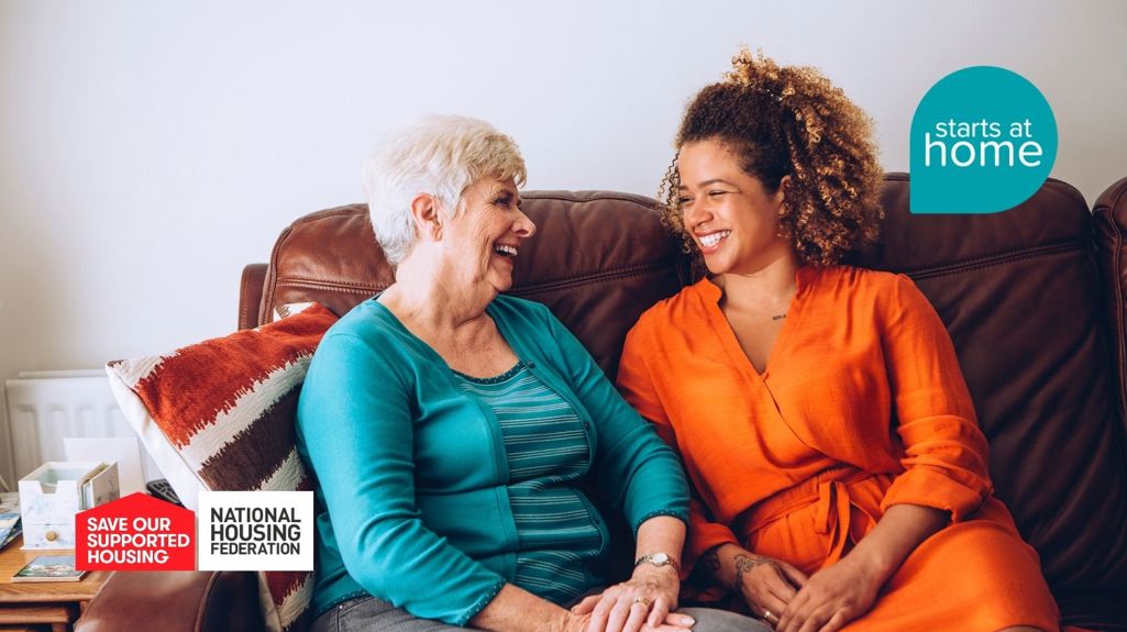 Two women sitting on a sofa, smiling warmly at each other and holding hands. The image includes logos for 'Save Our Supported Housing,' 'National Housing Federation,' and 'Starts at Home'."