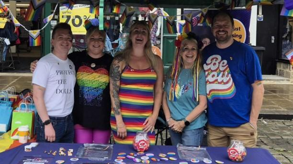 Five people stand behind a table at a Pride event stall decorated with rainbow flags and bunting. The table is covered with a purple cloth displaying the “Yorkshire Housing” logo and has flyers, badges, and small giveaways laid out. 