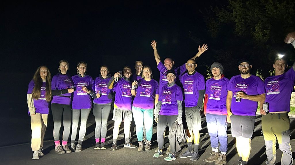 A group of colleagues all wearing matching purple t-shirts at the finish line of the Yorkshire Three Peaks, it's pretty dark so they're lit up by car headlights!