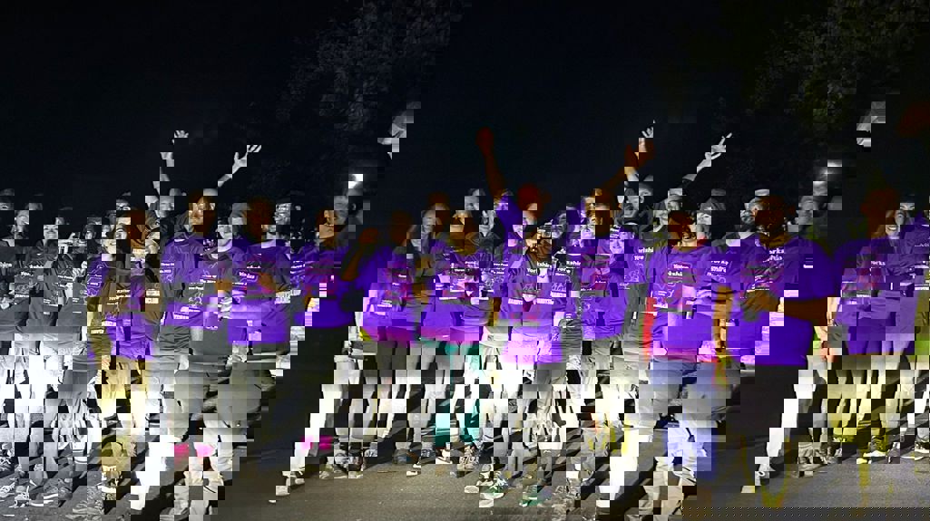 A group of colleagues all wearing matching purple t-shirts at the finish line of the Yorkshire Three Peaks, it's pretty dark so they're lit up by car headlights!