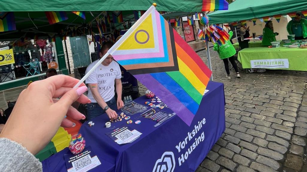 A hand holds a Progress Pride flag in the foreground at an outdoor community stall event, with green pop-up tents decorated with rainbow bunting behind it. A table covered with a purple “Yorkshire Housing” cloth displays badges and leaflets on a cobblestone street, with other charity stalls and people in the background beneath an overcast sky.