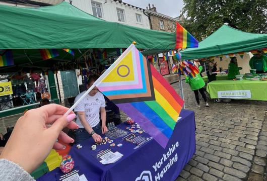 A hand holds a Progress Pride flag in the foreground at an outdoor community stall event, with green pop-up tents decorated with rainbow bunting behind it. A table covered with a purple “Yorkshire Housing” cloth displays badges and leaflets on a cobblestone street, with other charity stalls and people in the background beneath an overcast sky.