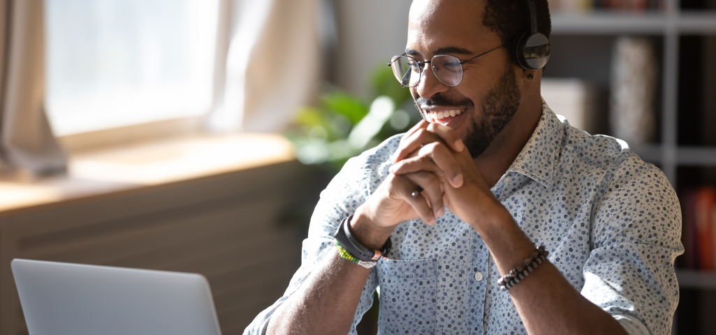 Man wearing a patterned shirt and glasses smiling at his computer, wearing a headset. 