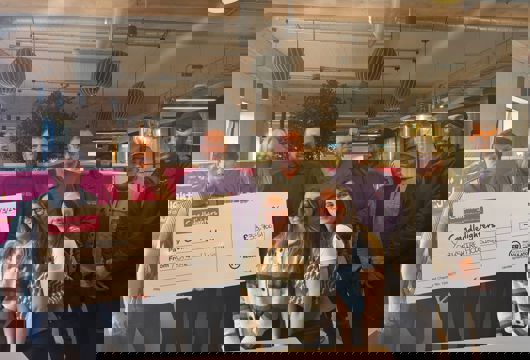 A group of nine people standing indoors in a modern office space, holding a large presentation cheque. The cheque reads “Candlelighters” and “From Yorkshire Housing” with an amount of £33,700.51 and a date of 31/3/26. Hanging lights, exposed ceiling beams, and plants are visible in the background.