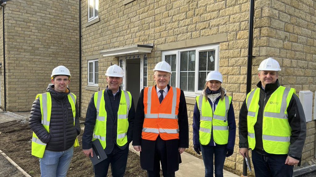 Group of four men and one woman wearing hi-vis jackets and hard hats standing outside a new house